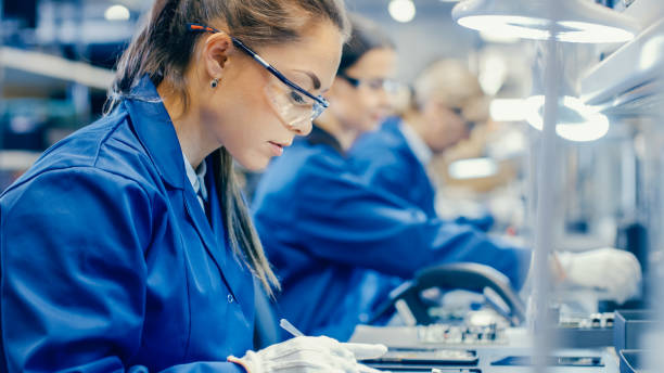 Female Electronics Factory Workers in Blue Work Coat and Protective Glasses Assembling Printed Circuit Boards for Smartphones with Tweezers. High Tech Factory with more Employees in the Background.